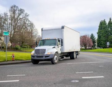 Small compact bonnet white semi truck rig with cube box trailer transporting commercial cargo driving on the street of urban city in spring time with blooming trees"n